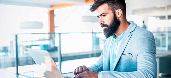 karriere tutor Ein bärtiger Mann in einem hellblauen Blazer arbeitet in einem modernen, hellen Büroraum an einem Laptop. Er wirkt konzentriert, vielleicht recherchiert er gerade Kursnet Bildungsangebote oder AZAV-zertifizierte Weiterbildung. Er sitzt an einem Schreibtisch mit einem großen Fenster im Hintergrund und genießt reichlich natürliches Licht und Inspiration.