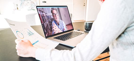 karriere tutor Eine Person sitzt an einem Tisch und hält ein Dokument mit Grafiken und Daten in der Hand, während sie an einer Online-Weiterbildung mit Bildungsgutschein auf einem Laptop teilnimmt. Auf dem Bildschirm ist eine Person mit Brille zu sehen, die in einem Raum mit Regalen im Hintergrund sitzt. Neben dem Laptop steht eine schwarze Tasse.