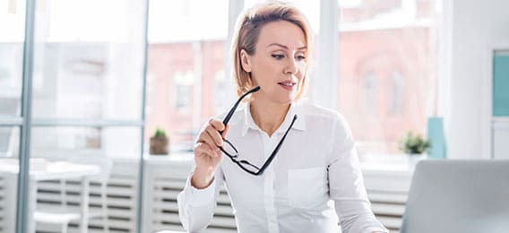 karriere tutor Eine Person mit kurzen blonden Haaren und weißem Hemd sitzt an einem Schreibtisch und hält eine Brille in der Hand. Sie wirkt konzentriert, während sie auf ihrem Laptop-Bildschirm die Kursnet-Bildungsangebote durchsucht. Im Hintergrund ist ein heller Raum mit großen Fenstern und unscharfer Außenlandschaft zu sehen.