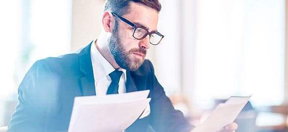 karriere tutor Ein Mann in Anzug und Brille sitzt an einem Schreibtisch und liest konzentriert Dokumente zur AZAV-zertifizierten Weiterbildung. Der Hintergrund ist leicht verschwommen, was auf eine Büroumgebung schließen lässt. Er wirkt konzentriert und in Gedanken versunken.