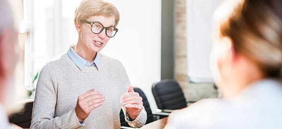 karriere tutor Eine Person mit kurzen blonden Haaren, Brille und hellem Pullover spricht angeregt über die geförderte Weiterbildung und gestikuliert dabei mit den Händen. Zwei weitere Personen sind leicht unscharf im Vordergrund zu sehen und hören in einem hellen, sonnendurchfluteten Raum aufmerksam zu.