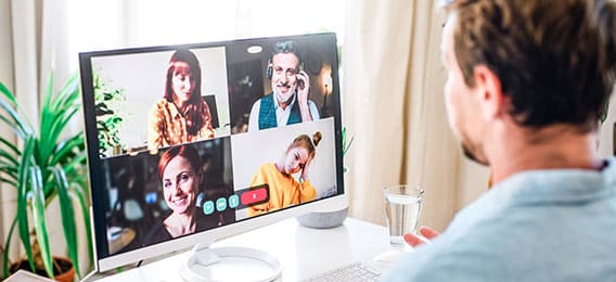 karriere tutor Ein Mann nimmt von seinem Homeoffice aus an einer Videokonferenz teil. Er sitzt an einem Schreibtisch mit einem Glas Wasser und einem Computerbildschirm, auf dem vier Kollegen im Gespräch zu sehen sind. Im Hintergrund sind Zimmerpflanzen und Sonnenlicht zu sehen, während er über eine von der Agentur für Arbeit finanzierte AZAV-zertifizierte Weiterbildung spricht.