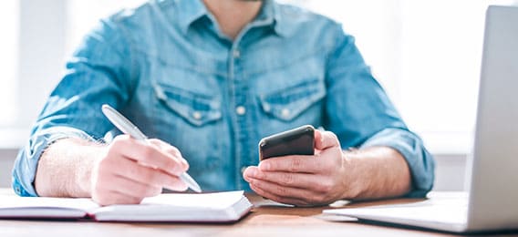 karriere tutor Eine Person im Jeanshemd sitzt mit einem aufgeschlagenen Notebook an einem Schreibtisch, hält in der einen Hand einen Stift, in der anderen ein Smartphone und recherchiert möglicherweise zu einer AZAV-zertifizierten Weiterbildung. Auf der rechten Bildseite ist teilweise ein Laptop zu sehen. Der Hintergrund ist sanft durch Tageslicht beleuchtet.